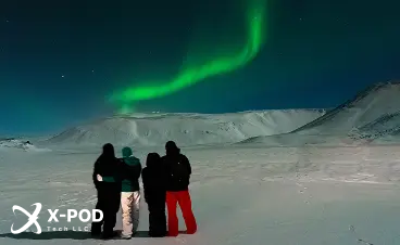 Three people watching the northern lights over snowy mountains at night.